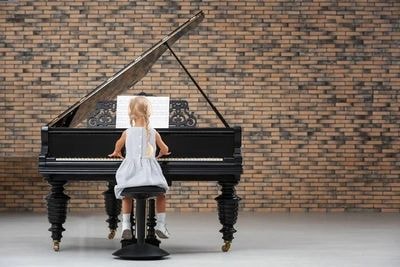 A young girl playing a grand piano against a brick wall.